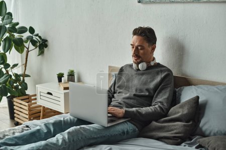 bärtiger Mann mit drahtlosen Kopfhörern per Laptop im Bett sitzend, freiberufliches Konzept