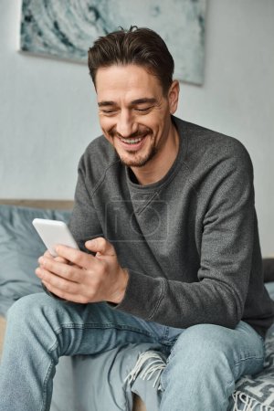 cheerful bearded man in grey sweater texting on smartphone in modern bedroom, social media