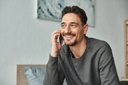cheerful bearded man in grey sweater having phone call on smartphone in modern bedroom, device