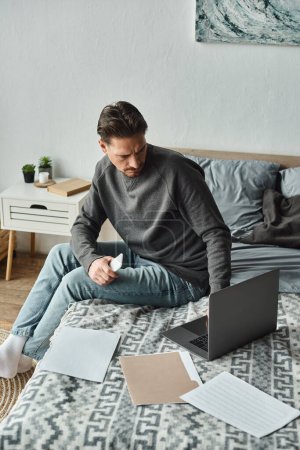 konzentrierter bärtiger Mann mit Smartphone und Laptop in der Nähe von Dokumenten auf dem Bett, Fernarbeit