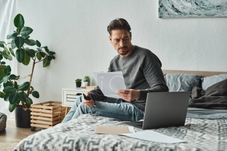 fokussierter und bärtiger Mann mit Smartphone und Laptop in der Nähe von Dokumenten auf dem Bett, Fernarbeit