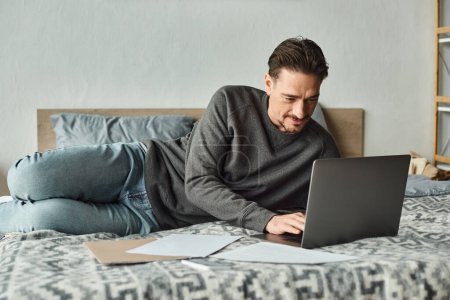 fokussierter und bärtiger Mann mit Laptop, während er von zu Hause aus im Schlafzimmer arbeitet, Dokumente