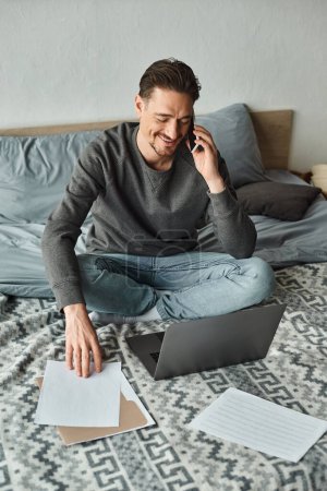 fröhlicher und bärtiger Mann, der Laptop und Smartphone benutzt, während er von zu Hause aus im Schlafzimmer arbeitet