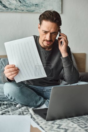 glücklicher und bärtiger Mann mit Laptop und Smartphone, während er von zu Hause aus im Schlafzimmer arbeitet