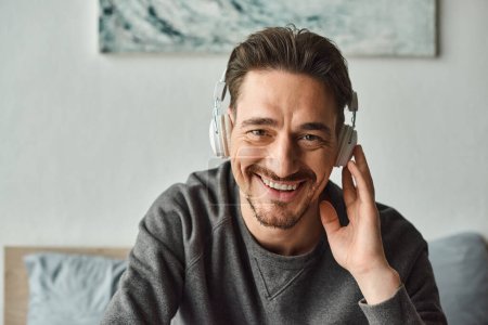 cheerful man in wireless headphones and grey casual sweater listening music and looking at camera