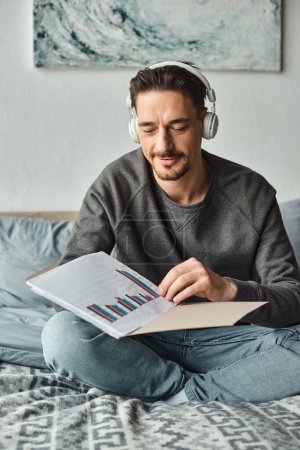 bärtiger Mann mit drahtlosen Kopfhörern, der Grafiken analysiert, während er im Schlafzimmer Musik hört, von zu Hause aus arbeitet