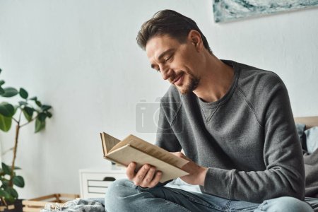 zufriedener Mann im grauen lässigen Pullover, der Buch liest, während er am Wochenende im Schlafzimmer entspannen kann, Freizeit