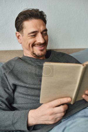 fröhlicher Mann im grauen lässigen Pullover, der Buch liest, während er am Wochenende im Schlafzimmer entspannen kann, Freizeit