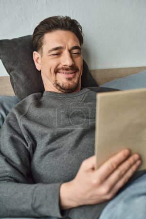 fröhlicher Mann im lässigen Pullover liest Buch und entspannt am Wochenende im Schlafzimmer, Freizeitkonzept