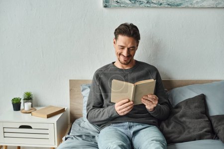 fröhlicher Mann im gemütlichen Pullover liest Buch und entspannt am Wochenende im Schlafzimmer, Freizeitkonzept