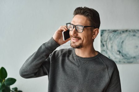 Photo for Bearded man in eyeglasses and grey jumper talking on smartphone in modern bedroom, phone call - Royalty Free Image