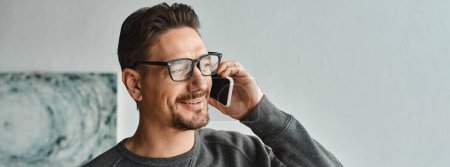 Photo for Bearded man in eyeglasses and grey jumper talking on smartphone in bedroom, phone call banner - Royalty Free Image