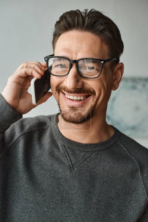 fröhlicher Mann mit Brille und grauem Pullover, der im modernen Schlafzimmer mit dem Smartphone telefoniert