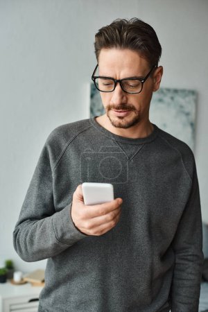 bärtiger Mann mit Brille und grauem Pullover, der im Schlafzimmer auf sein Smartphone schaut, Social-Media-Konzept