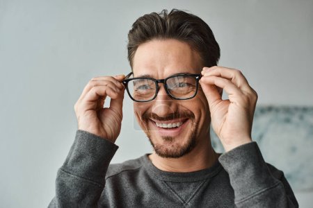 Photo for Portrait of cheerful and bearded man in grey jumper wearing eyeglasses and looking at camera - Royalty Free Image