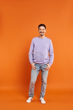 full length of cheerful and bearded man in purple sweater looking at camera on orange background