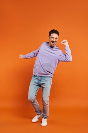 volle Länge des aufgeregten und bärtigen Mannes in lila Pullover und Jeans, der auf orangefarbenem Hintergrund tanzt