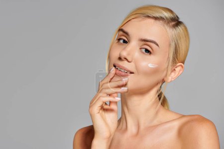 pretty girl with clear skin and cream on cheek touching chin with hand in gray background