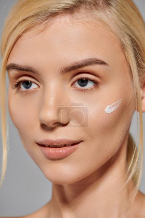 alluring young woman face with perfect glowing skin and cream on cheek against gray background