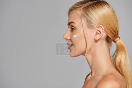 profile charming girl in her 20s with healthy skin and cream on cheek against grey background
