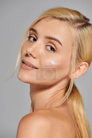 beautiful young woman with lotion on her cheek looking over her shoulder against grey background