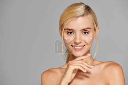 alluring girl in her 20s with cream on cheeks touching her chin against gray background