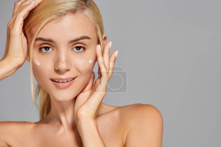 portrait charming blonde girl with her hands around head and cream on cheeks against grey background
