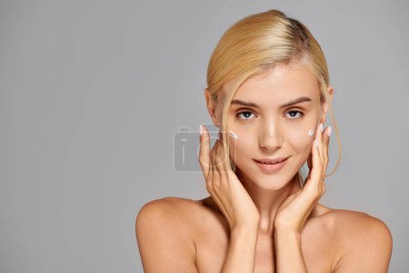 confident girl with blonde hair and cream on cheeks frames her neck with hands in grey background