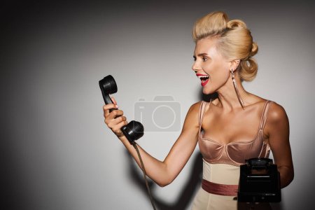 glamourous blonde woman in her 20s shouting angrily into retro phone against grey background