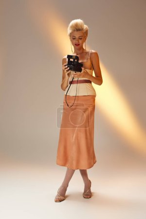 Charmante junge Frau mit blonden Haaren posiert mit Retro-Kamera vor grauem Hintergrund
