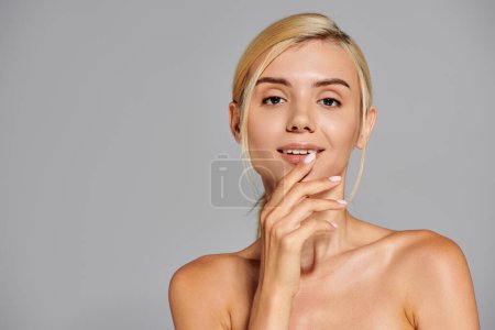 charming blonde girl in her 20s touching her lips with hand against gray background