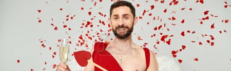 cheerful bearded man dressed as cupid with arrow and champagne under red confetti on grey, banner