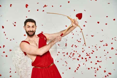 excitado barbudo hombre Cupido arqueo bajo confeti en forma de corazón en gris, San Valentín fiesta
