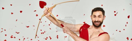 Foto de Divertido barbudo hombre Cupido arqueo bajo confeti rojo durante San Valentín partido en gris, bandera - Imagen libre de derechos