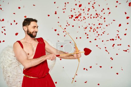 sonriente hombre Cupido barbudo arqueando con flecha en forma de corazón bajo confeti rojo, día de San Valentín
