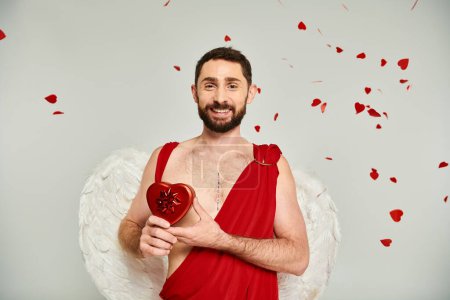 happy bearded man dressed as cupid with red hear-shaped st valentines present on grey, costume party