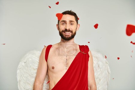bearded man in cupid costume and wings smiling at camera under red heart-shaped confetti on grey
