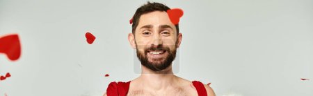 gracioso hombre Cupido sonriendo a la cámara bajo confeti rojo en forma de corazón, fiesta de San Valentín, pancarta