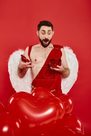 amazed man in cupid costume opening st valentines day present near heart-shaped balloons on red