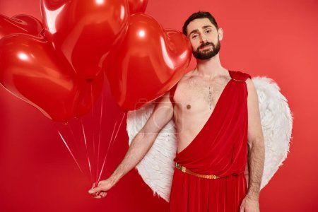 hombre de Cupido soñador y feliz con globos en forma de corazón mirando a la cámara en rojo, San Valentín día
