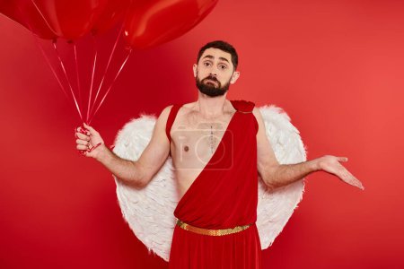 hombre barbudo en traje de Cupido con globos en forma de corazón mostrando gesto encogiéndose de hombros sobre fondo rojo