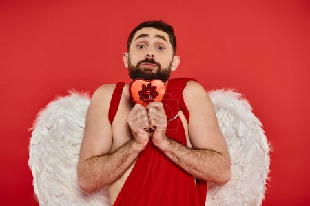 lustiger Mann in Amor-Kostüm und Flügeln mit herzförmigem Geschenkkarton und Blick in die Kamera auf rot