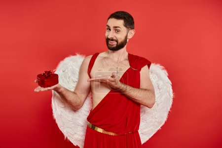 fröhlicher bärtiger Mann im Amor-Kostüm in herzförmiger Geschenkschachtel auf rot, Geschenk zum Valentinstag