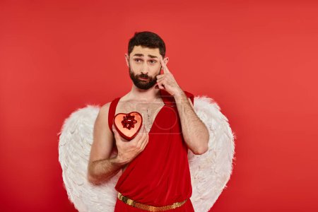 thoughtful bearded man in cupid costume with heart-shaped gift box pointing at his head on red