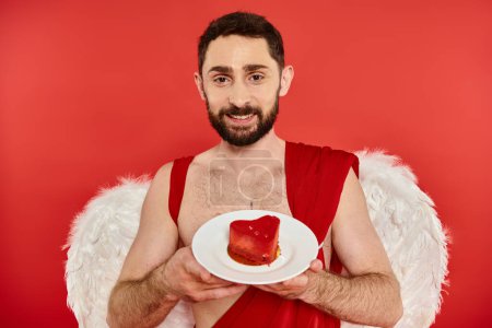 glücklicher bärtiger Mann im Amor-Kostüm mit leckerem herzförmigen Kuchen und Blick in die Kamera auf rot