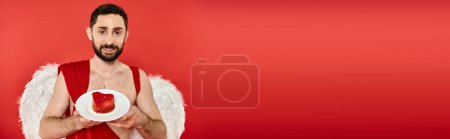 smiling man dressed as cupid showing delicious heart-shaped st valentines day cake on red, banner