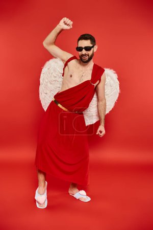 longitud completa de hombre excitado en traje de Cupido y gafas de sol bailando en rojo, fiesta de San Valentín