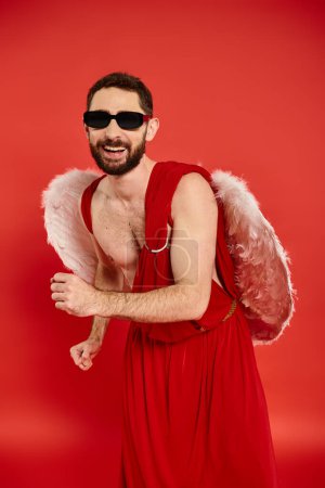 Schrulliger bärtiger Mann mit Sonnenbrille und Amor-Kostüm tanzt auf roter, Valentinstag-Motto-Party