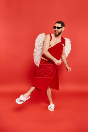 full length of excited bearded man in cupid costume dancing on red, st valentines day celebration