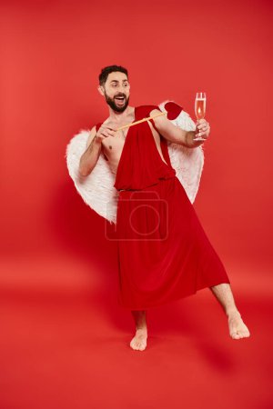 aufgeregter bärtiger Mann im Amor-Kostüm mit herzförmigem Pfeil und Champagnerglas auf rot, in voller Länge